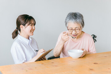 Wall Mural - 老人ホーム・家で食事する笑顔の高齢者女性と食事介助する管理栄養士・介護士・ヘルパーのアジア人女性
