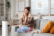 Frozen Young Woman With Cup Of Hot Tea Warming Near Electric Heater At Home