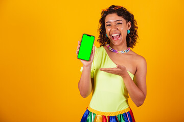 beautiful afro american brazilian woman, in carnival clothes, holding smartphone with green screen in chroma.