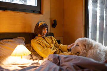 Canvas Print - Young woman cares her huge adorable white dog while lying in bed in tiny bedroom of wooden cabin on nature. House coziness and friendship with pets concept