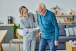 © AnnaStills - Young caregiver in uniform helping senior man to walk along the room, she working in nursing home