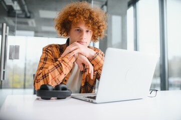 Portrait of Caucasian male freelancer in trendy apparel sitting at cafeteria table and doing remote work for programming design of public website, skilled software developer posing in coworking space.