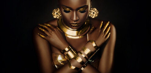 Art portrait african woman fashion model on black background, gold luxury accessories bijouterie jewelry bracelet earrings. Beautiful face sexy girl metallic golden professional makeup shiny lips skin