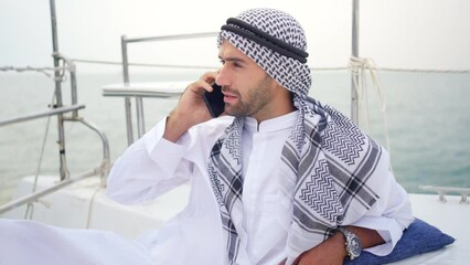 Wall Mural - Arab businessman talking on mobile phone with global corporate business while travel on private catamaran boat yacht sailing in the ocean. Arabic man enjoy luxury outdoor lifestyle on summer vacation.