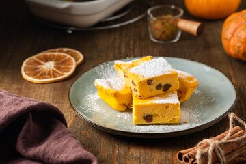 Canvas Print - Sliced cottage cheese casserole with pumpkin and raisins, sprinkled with powdered sugar, on a ceramic plate on a brown wooden background.