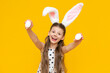 A little beautiful girl with masquerade ears of an Easter bunny on her head holds a chicken Easter egg in her hands and enjoys the Easter feast, on a yellow isolated background.
