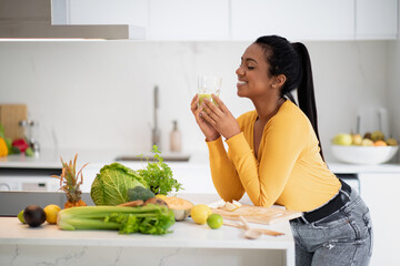 Wall Mural - Smiling young african american female drinking glass of smoothie at table with organic vegetables, enjoy diet