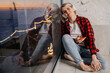 © Iona - Blonde American girl in casual plaid shirt and blue jeans sitting on windowsill using headphones, phone listening music enjoys sunset and city lights being in romantic moos. Dreamer, woman in love.