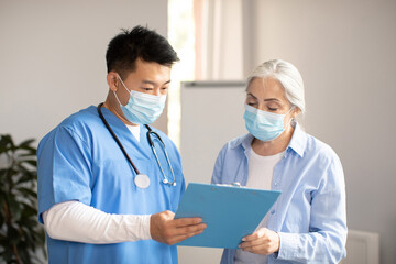 Wall Mural - Serious middle aged korean doctor in protective mask giving contract sign to old caucasian female patient