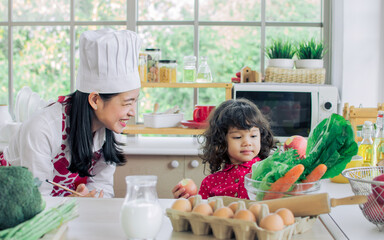 Beautiful adult mother wearing heart apron and hat, smiling at little daughter with love, fun, teaching for cooking together in kitchen, having breakfast at home. Education, Family Concept.