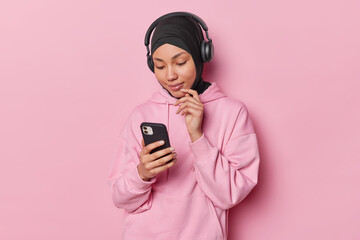 Horizontal shot of Muslim woman focused attentively at screen of smartphone chooses song to listen from playlist uses stereo headphones wears hijab and sweatshirt isolated over pink background