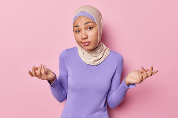 Canvas Print - Clueless woman spreads palms feels hesitant or questioned being doubtful wears hijab and purple turtleneck isolated over pink background doesnt know what to do. People and reactions concept.