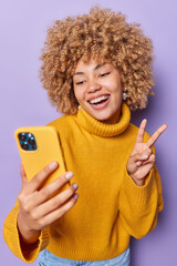 Wall Mural - positive beautiful woman with curly hair takes selfie via smartphone makes peace gesture smiles broa