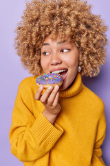 Wall Mural - Curly haired woman bites delicious glazed doughnut looks aside has sweet tooth wears yellow jumper with collar enjoys favorite dessert isolated over purple background. Unhealthy nutrition concept