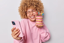 Overjoyed Curly Haired Woman Smiles Toothily Chuckles At Camera Holds Delicious Sweet Doughnuts Uses Mobile Phone For Taking Selfie Poses With Favorite Dessert Isolated Over Grey Background.