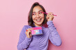 © wayhome.studio  - Happy Asian woman with dark hair applies pink eyeshadow uses cosmetic brush smiles broadly has white even teeth wears purple turtleneck isolated over pink background. Beauty and wellness concept
