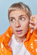 © WHstudio Leushin N - Headshot of surprised millennial girl with short blonde hair keeps hand raised near face looks wondered aside wears orange windbreaker feels impressed isolated over blue background. Close up shot