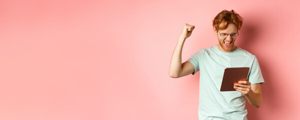 Image of happy redhead man triumphing, winning online with digital tablet and rejoicing, standing over pink background