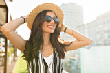 Fototapeta Boho - Close up outdoor photo of smiling happy woman with dark hair looking up and touching her hat while resting outdoor in the city 