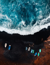 Vertical Aerial Photo Of Black Sand Volcanic Beach With Boats On The Shore And Big And Strong Waves In Ocean -  Lanzarote, Canary Islands. Waves In Blue Ocean From Above (top View) With Red Cliff.