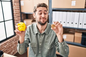 Wall Mural - Hispanic man with beard working at small business ecommerce holding piggy bank and bitcoin smiling looking to the side and staring away thinking.