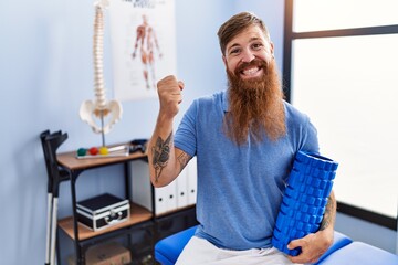 Poster - Redhead man with long beard holding foam roller at medical clinic screaming proud, celebrating victory and success very excited with raised arm