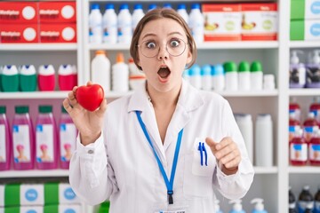 Sticker - Young caucasian woman working at pharmacy drugstore holding red heart scared and amazed with open mouth for surprise, disbelief face