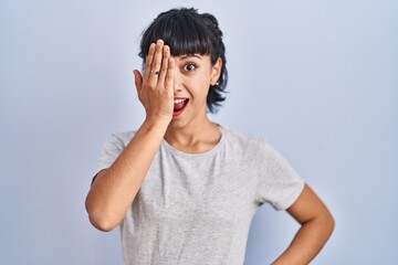 Wall Mural - Young hispanic woman wearing casual t shirt over blue background covering one eye with hand, confident smile on face and surprise emotion.