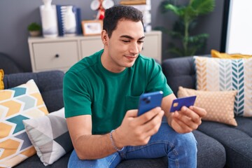 Sticker - Young hispanic man using smartphone and credit card sitting on sofa at home