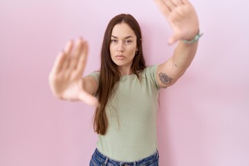 Poster - Beautiful brunette woman standing over pink background doing frame using hands palms and fingers, camera perspective