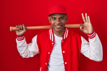 Poster - Young hispanic man playing baseball holding bat smiling positive doing ok sign with hand and fingers. successful expression.