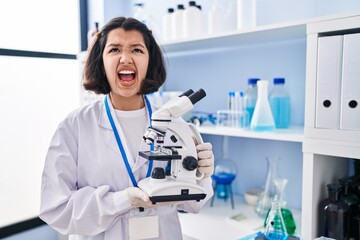 Sticker - Young hispanic woman working at scientist laboratory holding microscope angry and mad screaming frustrated and furious, shouting with anger looking up.