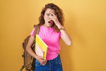 Poster - Young caucasian woman wearing student backpack and holding books peeking in shock covering face and eyes with hand, looking through fingers with embarrassed expression.