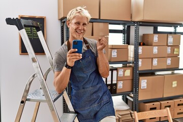 Wall Mural - Young blond man using smartphone working at storehouse very happy and excited doing winner gesture with arms raised, smiling and screaming for success. celebration concept.