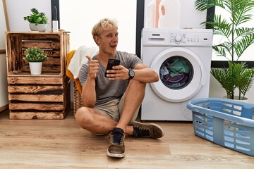 Wall Mural - Young blond man doing laundry using smartphone doing happy thumbs up gesture with hand. approving expression looking at the camera showing success.