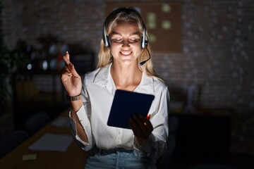 Poster - Young blonde woman working at the office at night gesturing finger crossed smiling with hope and eyes closed. luck and superstitious concept.