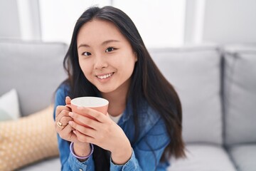 Canvas Print - Young chinese woman drinking coffee sitting on sofa at home