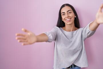 Wall Mural - Young brunette woman standing over pink background looking at the camera smiling with open arms for hug. cheerful expression embracing happiness.