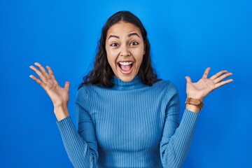 Wall Mural - Young brazilian woman standing over blue isolated background celebrating crazy and amazed for success with arms raised and open eyes screaming excited. winner concept