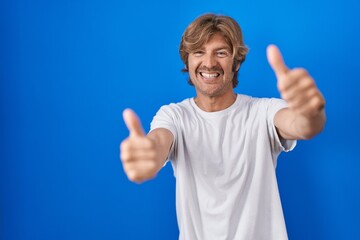 Poster - Middle age man standing over blue background approving doing positive gesture with hand, thumbs up smiling and happy for success. winner gesture.