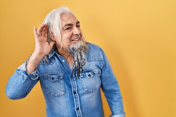 Poster - Middle age man with grey hair standing over yellow background smiling with hand over ear listening an hearing to rumor or gossip. deafness concept.