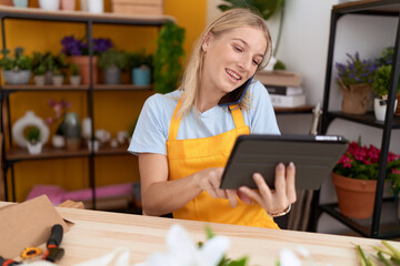 Wall Mural - Young blonde woman florist talking on smartphone using touchpad at flower shop