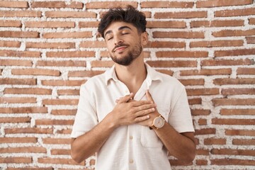 Poster - Arab man with beard standing over bricks wall background smiling with hands on chest with closed eyes and grateful gesture on face. health concept.