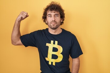 Canvas Print - Hispanic young man wearing bitcoin t shirt strong person showing arm muscle, confident and proud of power