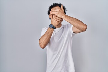 Poster - Hispanic man standing over isolated background covering eyes and mouth with hands, surprised and shocked. hiding emotion