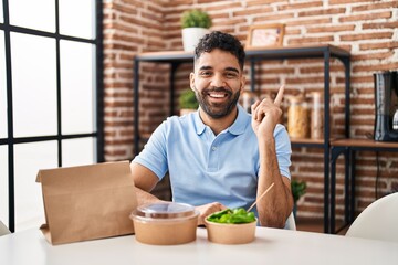 Wall Mural - Hispanic man with beard eating delivery salad smiling happy pointing with hand and finger to the side