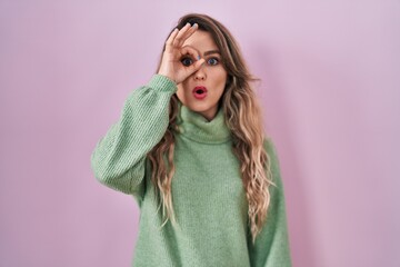 Canvas Print - Young caucasian woman standing over pink background doing ok gesture shocked with surprised face, eye looking through fingers. unbelieving expression.