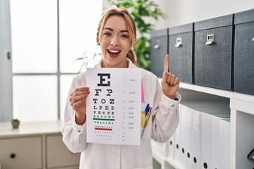 Poster - Hispanic optician woman holding medical exam smiling with an idea or question pointing finger with happy face, number one