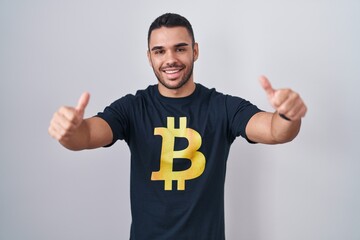 Canvas Print - Young hispanic man wearing bitcoin t shirt approving doing positive gesture with hand, thumbs up smiling and happy for success. winner gesture.