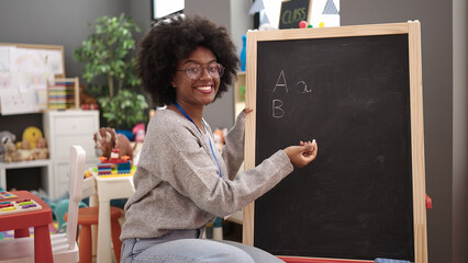 African american woman teacher smiling confident writing on blackboard at kindergarten